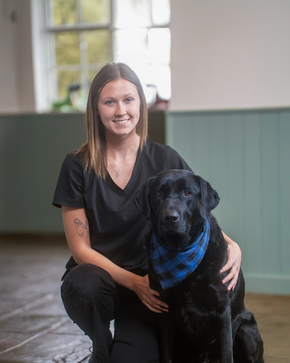 A young woman with shoulder-length brown hair, wearing a black scrub top and pants, kneels beside a black Labrador Retriever wearing a blue and black plaid bandana. The woman has a small tattoo on her forearm, is wearing a cross necklace, and has her hand resting on the dog's side. They are both facing forward against a backdrop of a pale green wall and a window.