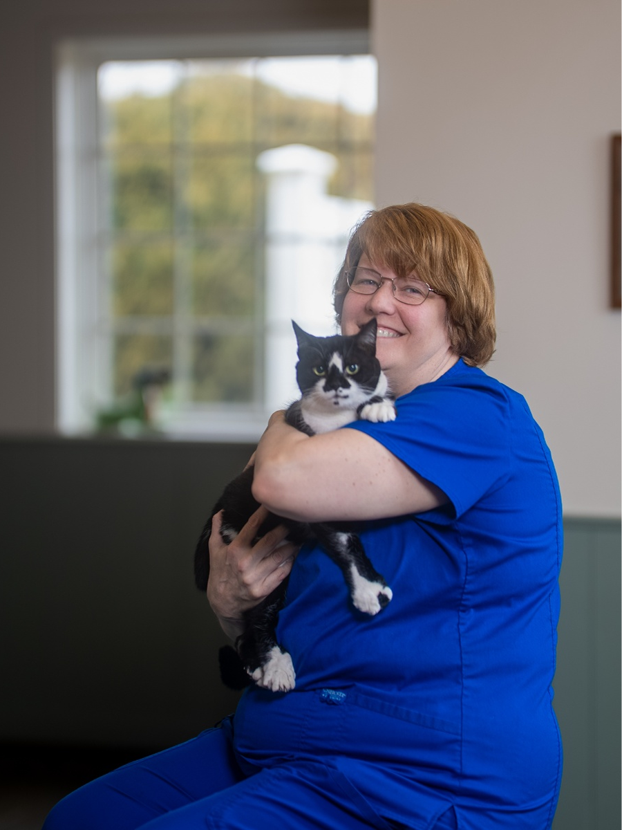 A woman with reddish-brown hair and glasses, wearing a blue medical scrub top, smiles while holding a black-and-white cat in her arms against an indoor background with a large window.