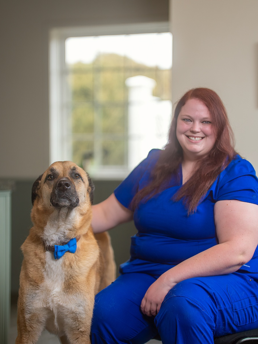 A woman with red hair wearing blue medical scrubs smiles while resting her hand on the back of a large, tan-colored dog wearing a blue bow tie.