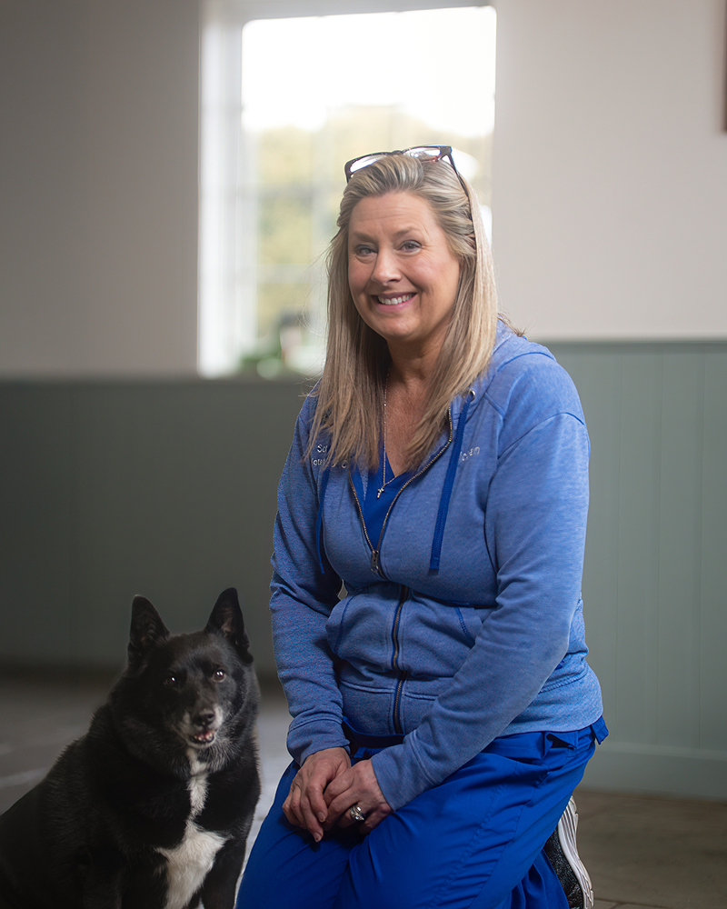 A blonde woman wearing a blue zip-up hoodie and blue scrubs kneeling on a light-colored floor next to a medium-sized black dog with a white chest patch, posing for a portrait in a room with a large window in the background.