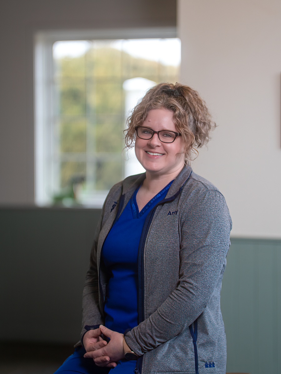 A smiling woman with curly light brown hair tied back, wearing glasses, a blue scrub top, and a grey zip-up jacket, poses for a portrait against a soft-focus indoor background with a large window.