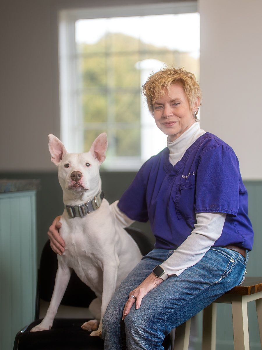 A blonde woman wearing a purple scrub top over a white turtleneck and blue jeans sits on a stool with her arm around a white dog with pointed ears and a grey collar.