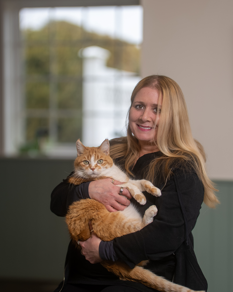 A woman with long blonde hair, wearing a black zip-up jacket, smiles while holding a large orange and white tabby cat in her arms.