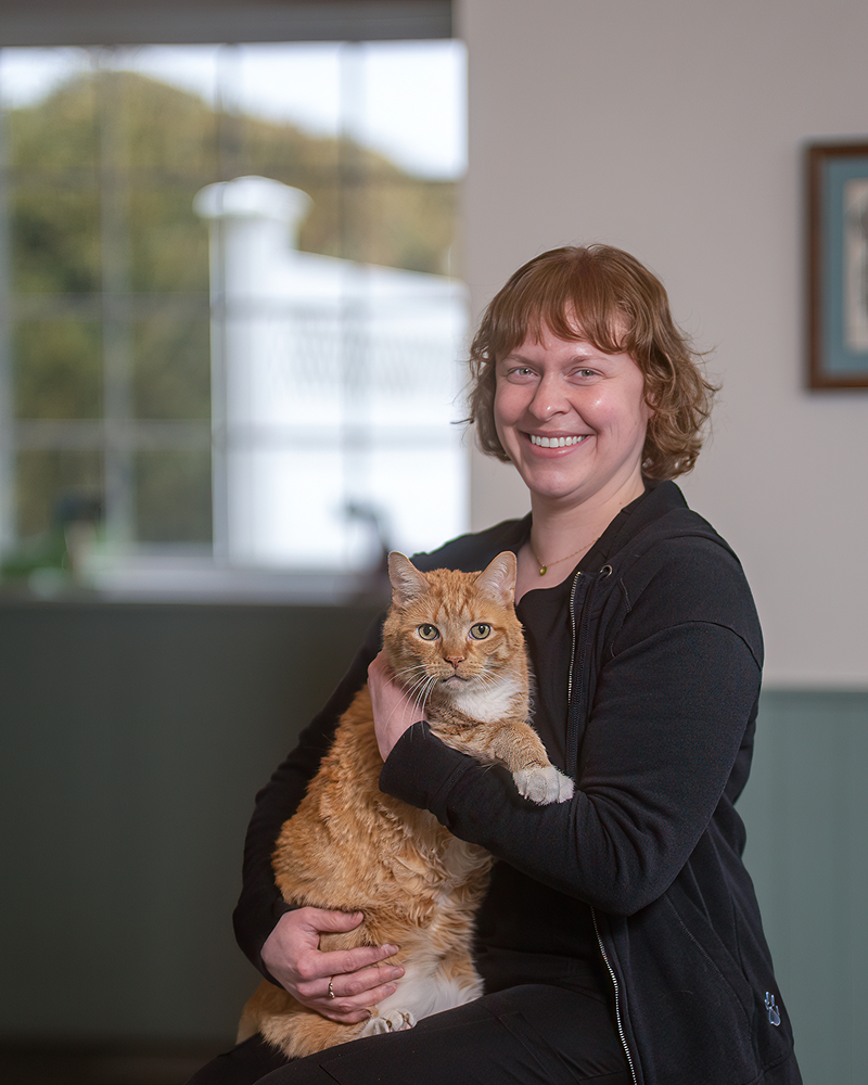 A woman with auburn hair and a friendly smile, dressed in a black zip-up hoodie, sits and holds a large orange tabby cat. The cat, which has white markings on its chest and paws, faces forward with a calm expression. The background features a large window with a blurred outdoor view and a framed picture on a neutral-colored wall.