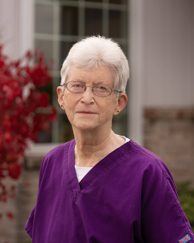 A portrait of an older woman with short, light hair and glasses, wearing a purple v-neck scrub top, standing outdoors in front of a window and a red-leaved tree.